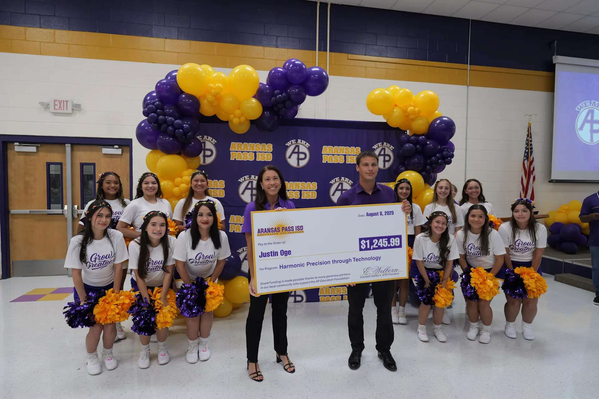 A teacher recieves an award from the Aransas Pass ISD Education Foundation. They are in front of a purple AP backdrop surrounded by the APHS Varsity Cheer Team