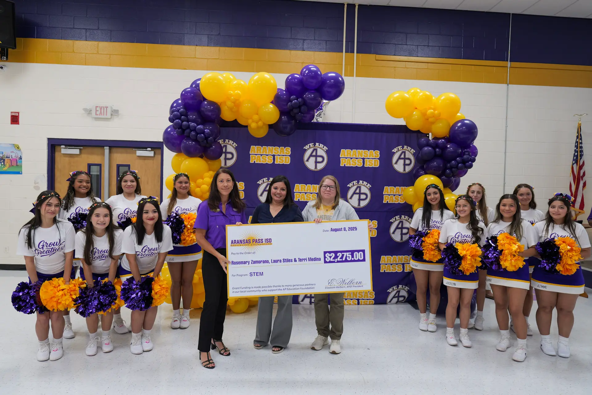 Two teachers are receiving an award from the APISD Education Foundation. The teachers are standing next to a Education Foundation member, and they are all surrounded by the APHS Varsity Cheerleaders