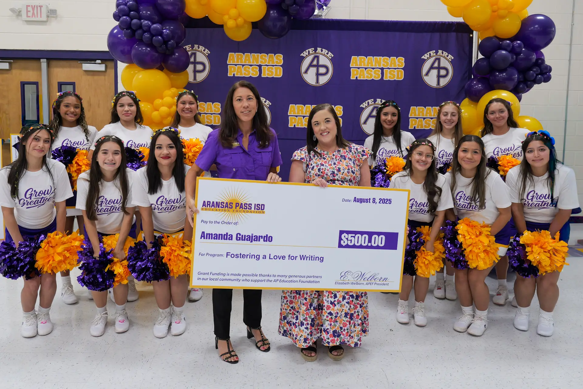 A teacher receives an award from the Aransas Pass ISD Education Foundation. The teach is being handed a check from a foundation member, and they are surrounded by the APHS Varsity Cheerleaders