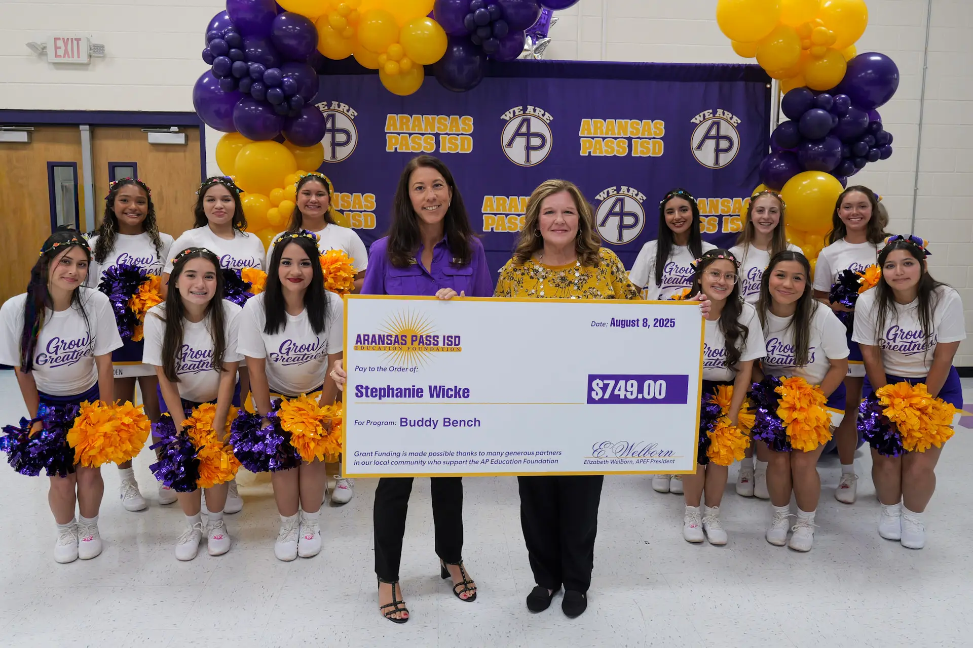 A teacher is accepting an award from the APISD Education Foundation. They are standing in front of a purple APISD backdrop and surrounded by the APHS Varsity cheerleaders