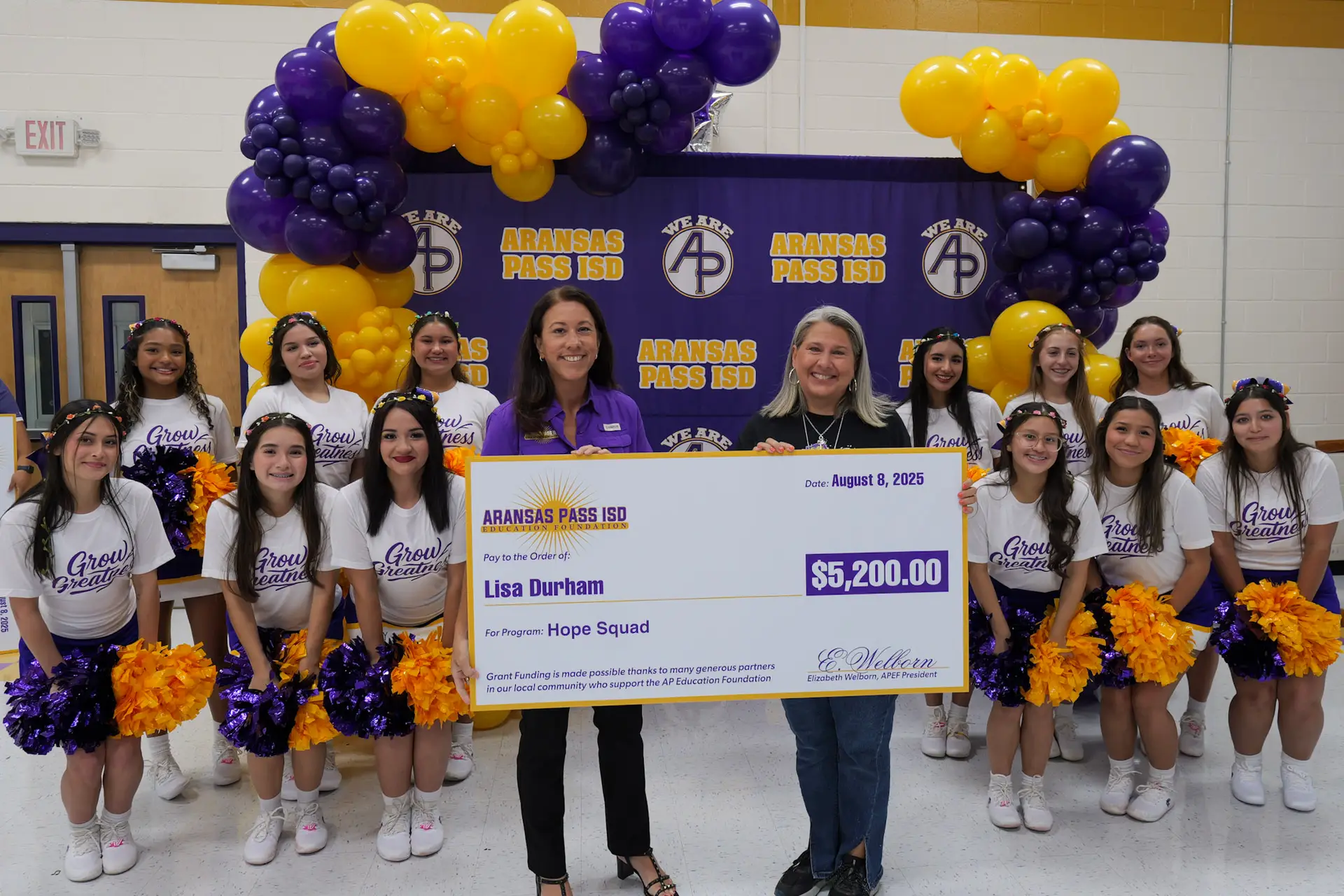 A teacher is being handed an award from the APISD Education Foundation. They are surrounded by the APISD Varsity Cheerleaders